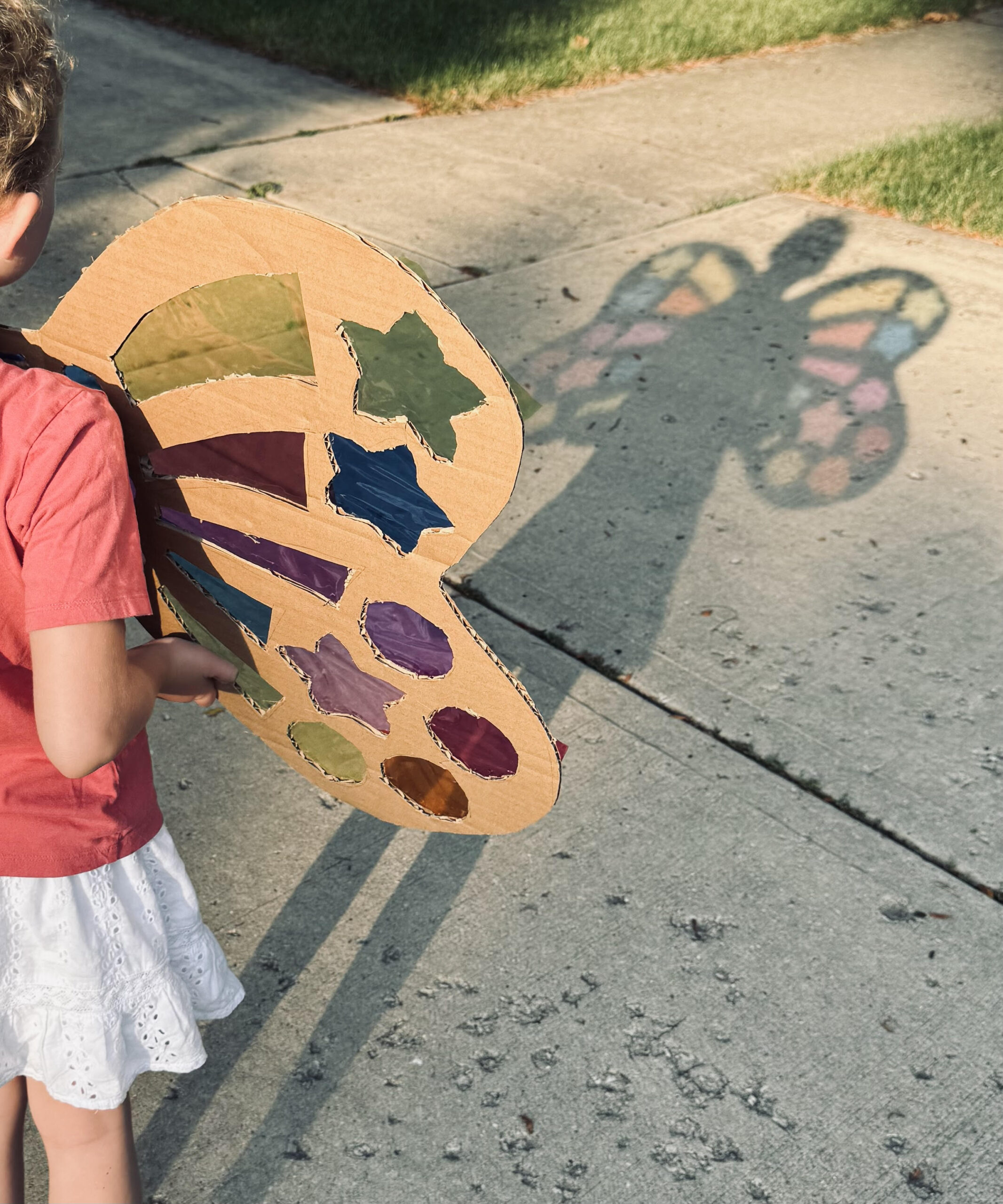 child holding handmade rainbow-colored fairy wings, sunlight reflecting colors on pavement, hands visible, playful creative scene.